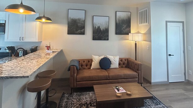 Uma sala de estar moderna com um sofá de couro marrom, uma mesa de centro e um tapete estampado. Há uma área de cozinha com bancada de mármore e iluminação pendente.