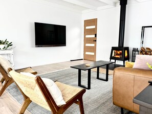 A modern living room with a flat-screen TV, a sofa, a wooden chair, a small table, and a wood-burning stove. - Little Red Gate Urban Retreat (Kingston)