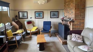 A living room with a brick fireplace, a blue armchair, a yellow armchair, a wooden coffee table, and a beige sofa. - The Cottage at Raheen (Mallacoota)