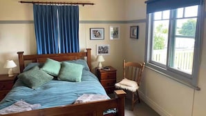 A bedroom with a wooden bed, blue curtains, a wooden chair, a nightstand with a lamp, and framed pictures on the wall. - The Cottage at Raheen (Mallacoota)