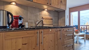 A kitchen with wooden cabinets, a stainless steel kettle, a red coffee maker, and a view of mountains through large windows. - Domaine des Reines D10 by Interhome (Nendaz)