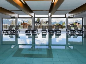 Ein großer Innenpool mit deutlichen Spiegelungen, umgeben von Liegestühlen und großen Fenstern mit Blick auf die Berge.