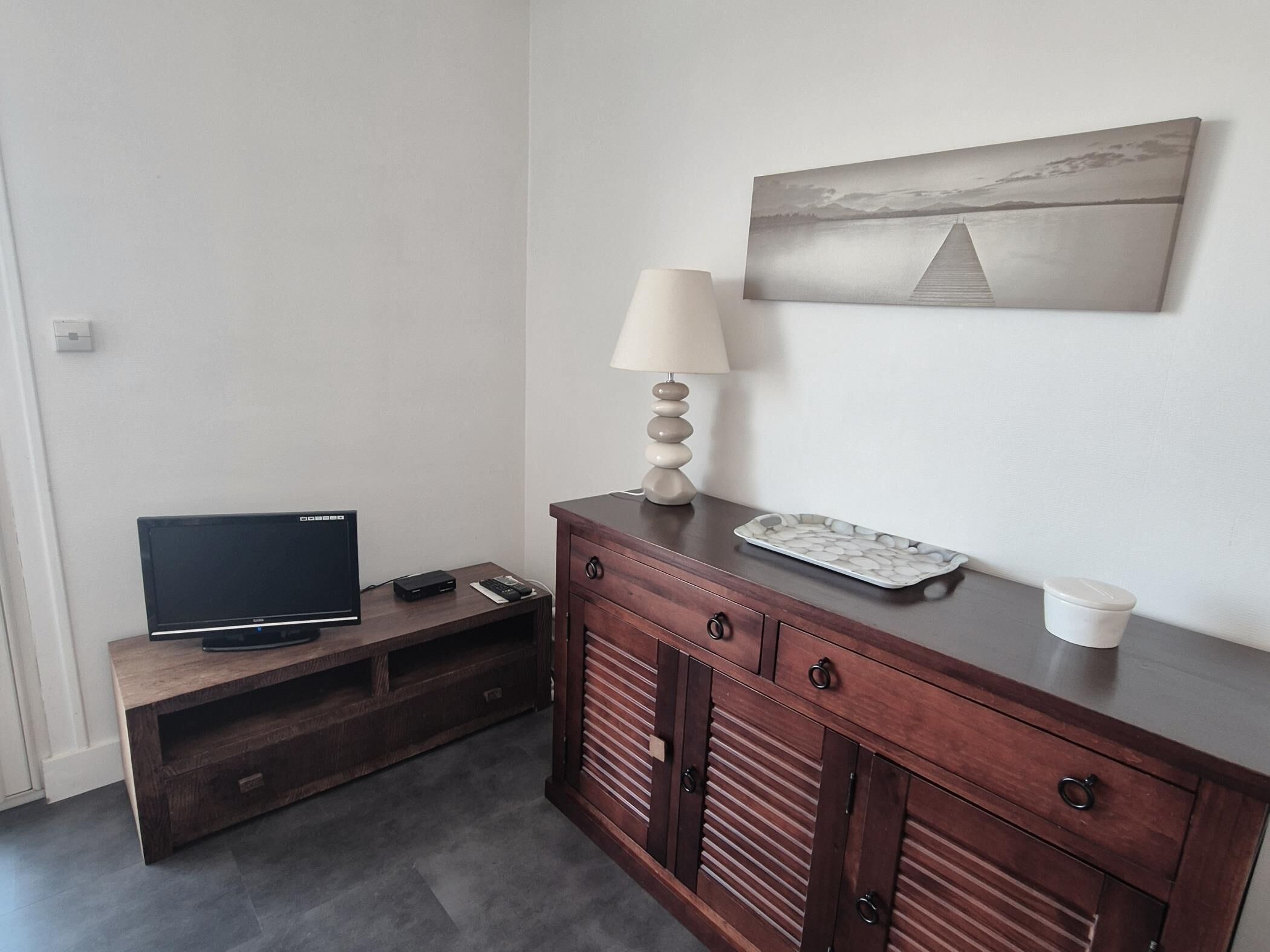 Une pièce avec un téléviseur posé sur un meuble en bois, une lampe, une petite table avec un plateau et un tableau accroché au mur.