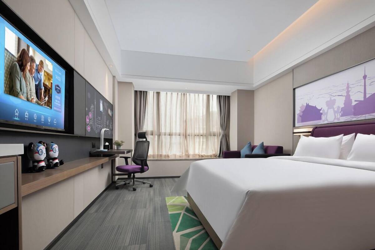 A modern hotel room with a large bed, a desk with a computer, a TV mounted on the wall, and a window with curtains.