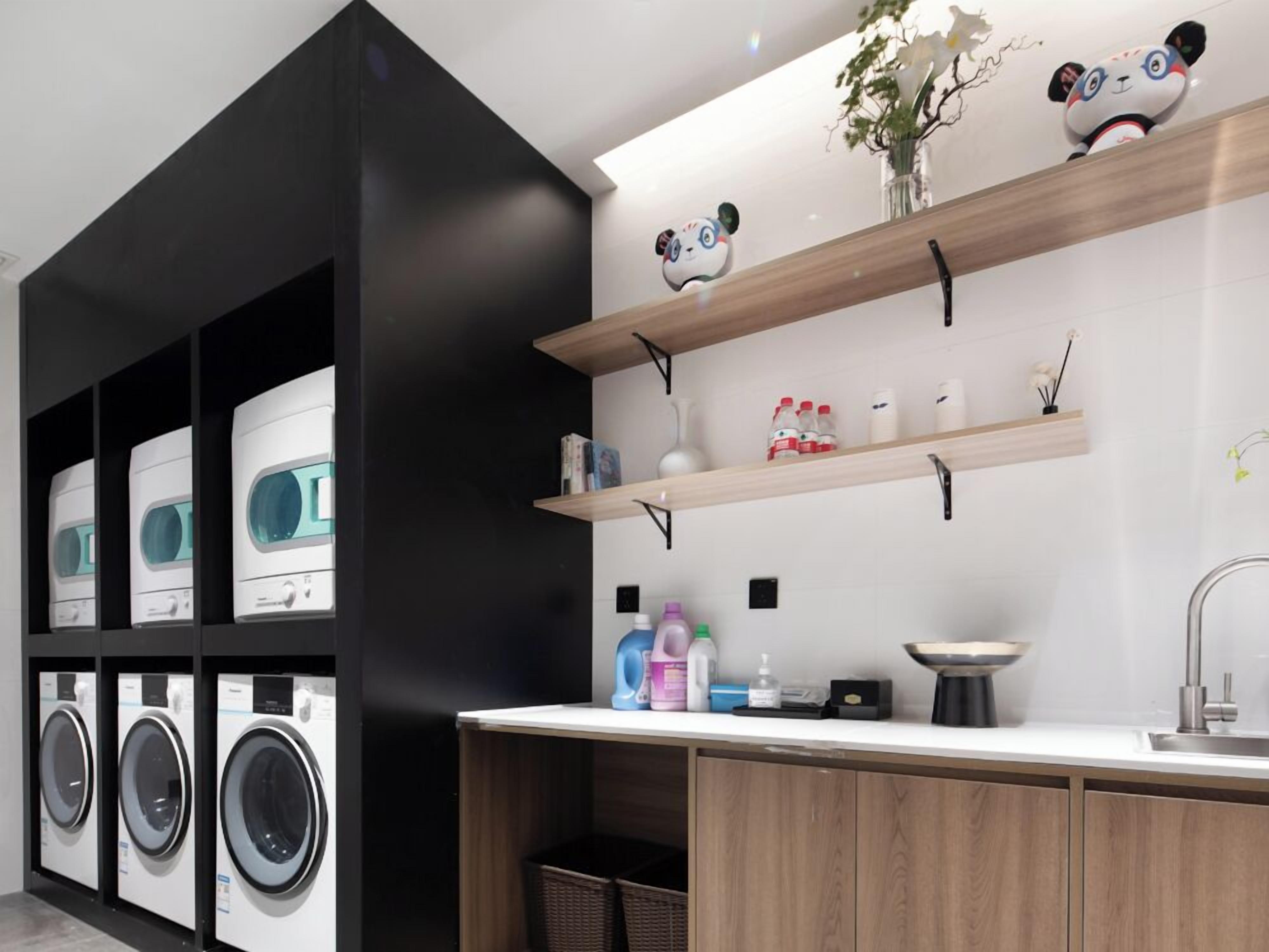 A modern laundry room with multiple washing machines, a sink, and wooden shelves with decorative items.