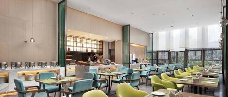 A spacious dining area with multiple tables and chairs, a kitchen area in the background, and large windows providing natural light.