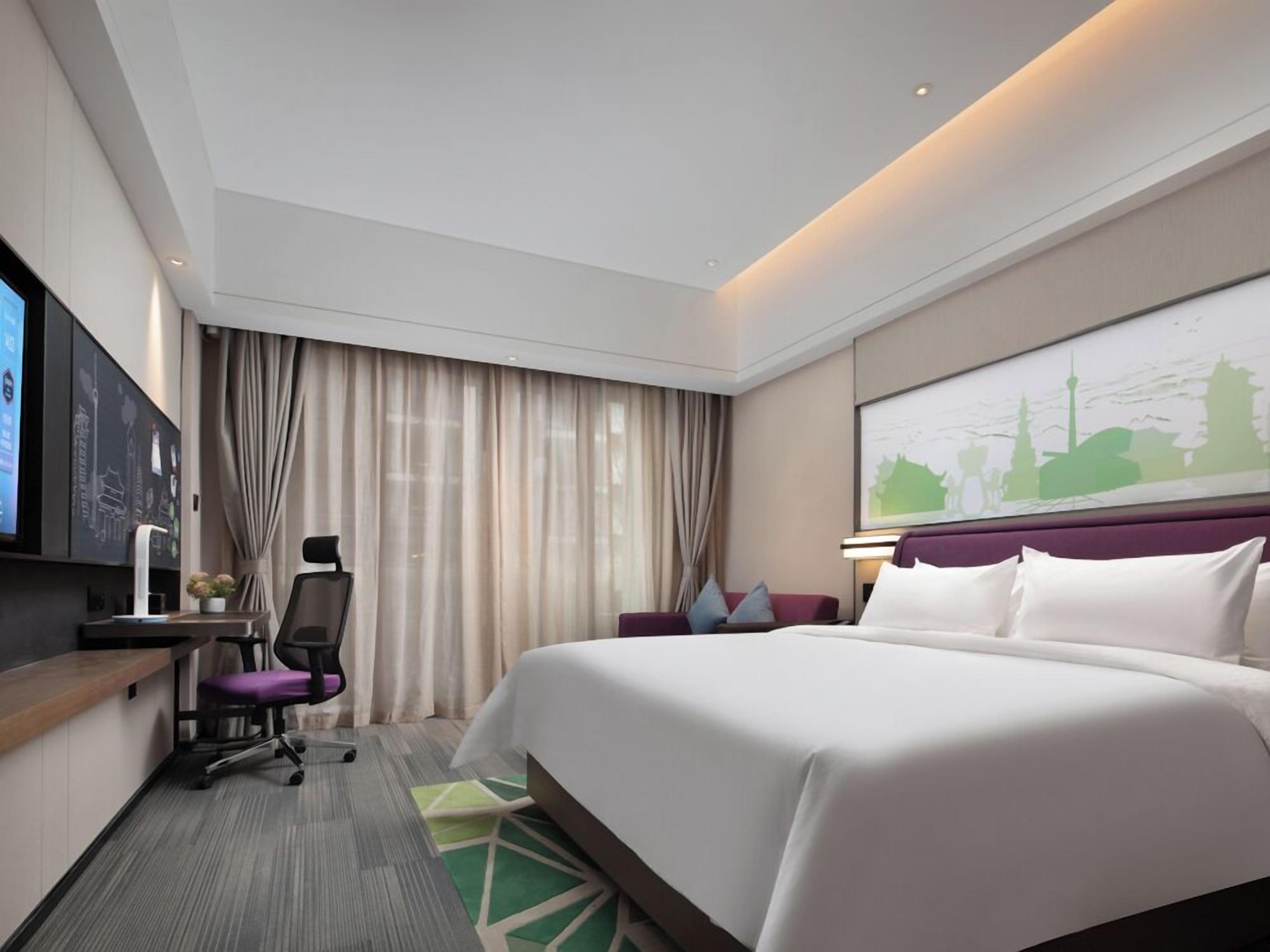 A modern hotel room with a large bed, a desk with a chair, a television, and a cityscape artwork on the wall.