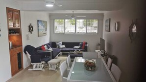 Ein modernes Wohnzimmer mit einer Couch, einem Esstisch und Stühlen. Es gibt Fenster mit Gitterstäben, einen Deckenventilator und einen Fernseher.
