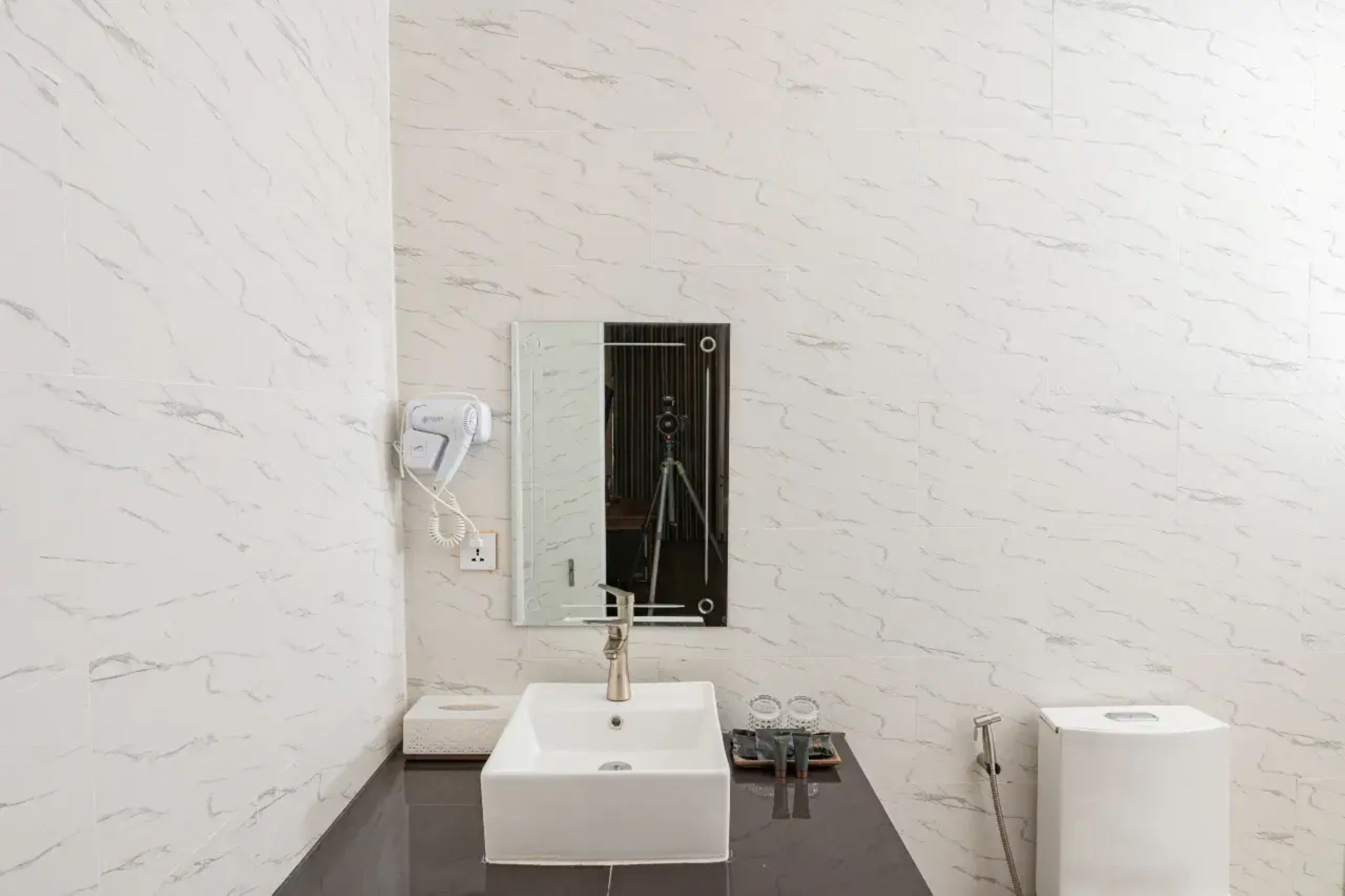 A modern bathroom with a white marble wall, a white sink, a mirror, a towel dispenser, and a shower unit.