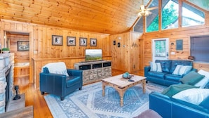 Interior acogedor de una cabaña con paredes de madera, una chimenea de piedra, dos sofás azules, una mesa de centro de madera y un ventilador de techo.