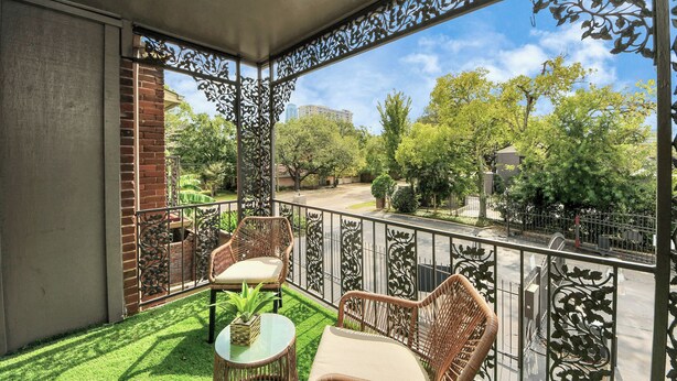 A balcony with wicker furniture, a small table, and a view of a garden and buildings. - Houston Galleria Suite 
Modern Rental 5 mins from the Galleria Mall (Houston)
