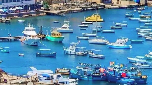 Un pueblo costero con una imponente estructura que recuerda a un castillo, un puerto deportivo con numerosos barcos y un mar azul cristalino.