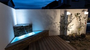 A nighttime outdoor area with a stone water feature, wooden deck, and illuminated plants.