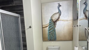 A bathroom with a white sink, a blue bath mat, a peacock painting on the wall, and a toilet.