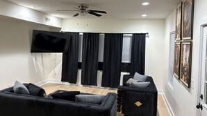 A modern living room with a flat-screen TV, black and grey sofas, a ceiling fan, and black curtains.