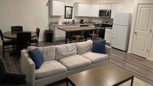 A modern kitchen with white cabinets, a microwave, and a refrigerator. There is a dining area with a table and chairs, and a living room with a sofa and a coffee table.