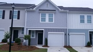A two-story residential building with a gray and white exterior, white garage doors, and a front yard with small shrubs and a street lamp.
