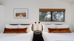A double bed room with white bedding and brown pillows, a bedside table with a lamp, a framed picture on the wall, and a window with a view of trees.