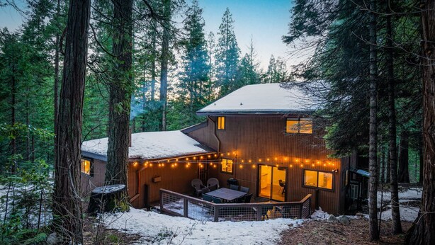 A cabin with a deck and string lights, surrounded by tall trees and snow.
