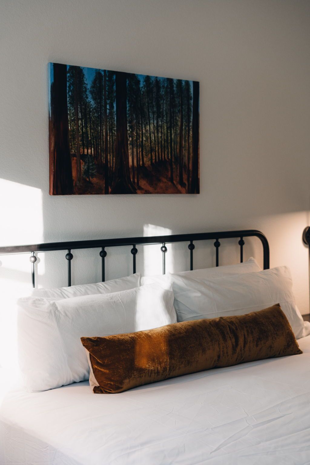 Un lit avec du linge de lit blanc et un oreiller marron, une tête de lit en métal noir et, au-dessus, un tableau sur le thème de la forêt.