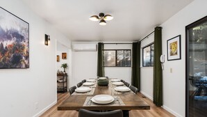 A dining area with a wooden table set for eight, surrounded by chairs, with a large painting on the left and a window with curtains on the right.