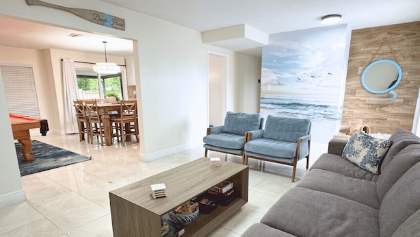 Un salon avec un canapé gris, des fauteuils bleus, une table basse en bois et une fresque murale sur le thème de la plage.