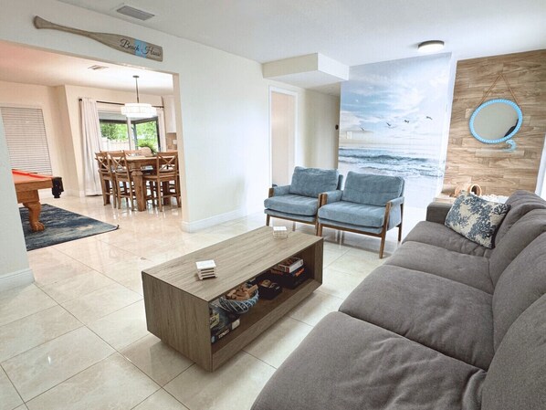 Ein Wohnzimmer mit grauem Sofa, blauen Sesseln, einem Holztisch und einem Wandbild mit Strandmotiv.