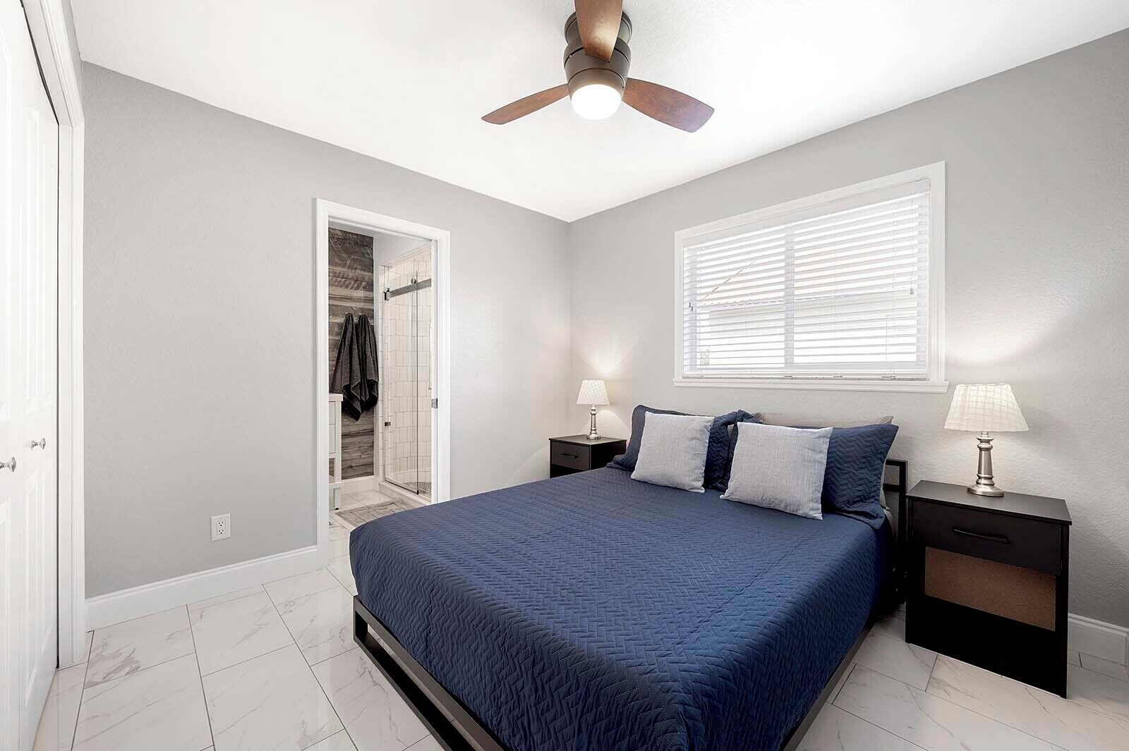 Une chambre à coucher comprenant un lit, des tables de chevet, un ventilateur de plafond, une fenêtre avec des stores et un dressing.