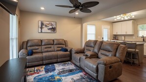 A living room with a brown leather sectional sofa, a ceiling fan, a colorful abstract rug, and a kitchen in the background. -  The Scholar's Retreat (Tuskegee)