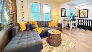 A living room with a grey sectional sofa, yellow cushions, a round wicker coffee table, and a patterned rug. There are two windows with blinds, a ceiling fan, and a dining area with a table and chairs. - Romantic Lakefront Getaway with Balcony near Downtown Orlando (Orlando)
