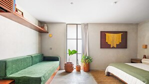 Een moderne slaapkamer met een groene bank, een bed met een witgroene dekbedovertrek, een groot raam met gordijnen en een hangplant aan de muur.