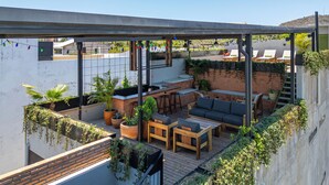 A rooftop patio with a wooden table, chairs, and a sofa. There are potted plants and a string of colorful lights. - ALMA Surf Lodge Terrace Queen with View (Corral del Risco)