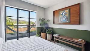 A bedroom with a large bed, a bench, a potted plant, a framed artwork, and a view of a hammock and greenery outside. - ALMA Surf Lodge Terrace Queen with View (Corral del Risco)