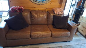 A brown leather sofa with two dark cushions, a woven wall hanging, and a wood-burning stove in a cabin-style room. - Hummingbird Heaven! Woodland Park Mountain Cabin (Woodland Park)