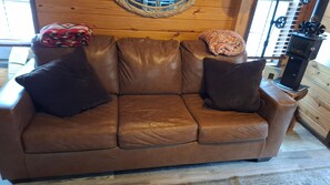 Ein braunes Ledersofa mit zwei dunklen Kissen, ein gewebter Wandbehang und ein Kaminofen in einem Blockhausszimmer.