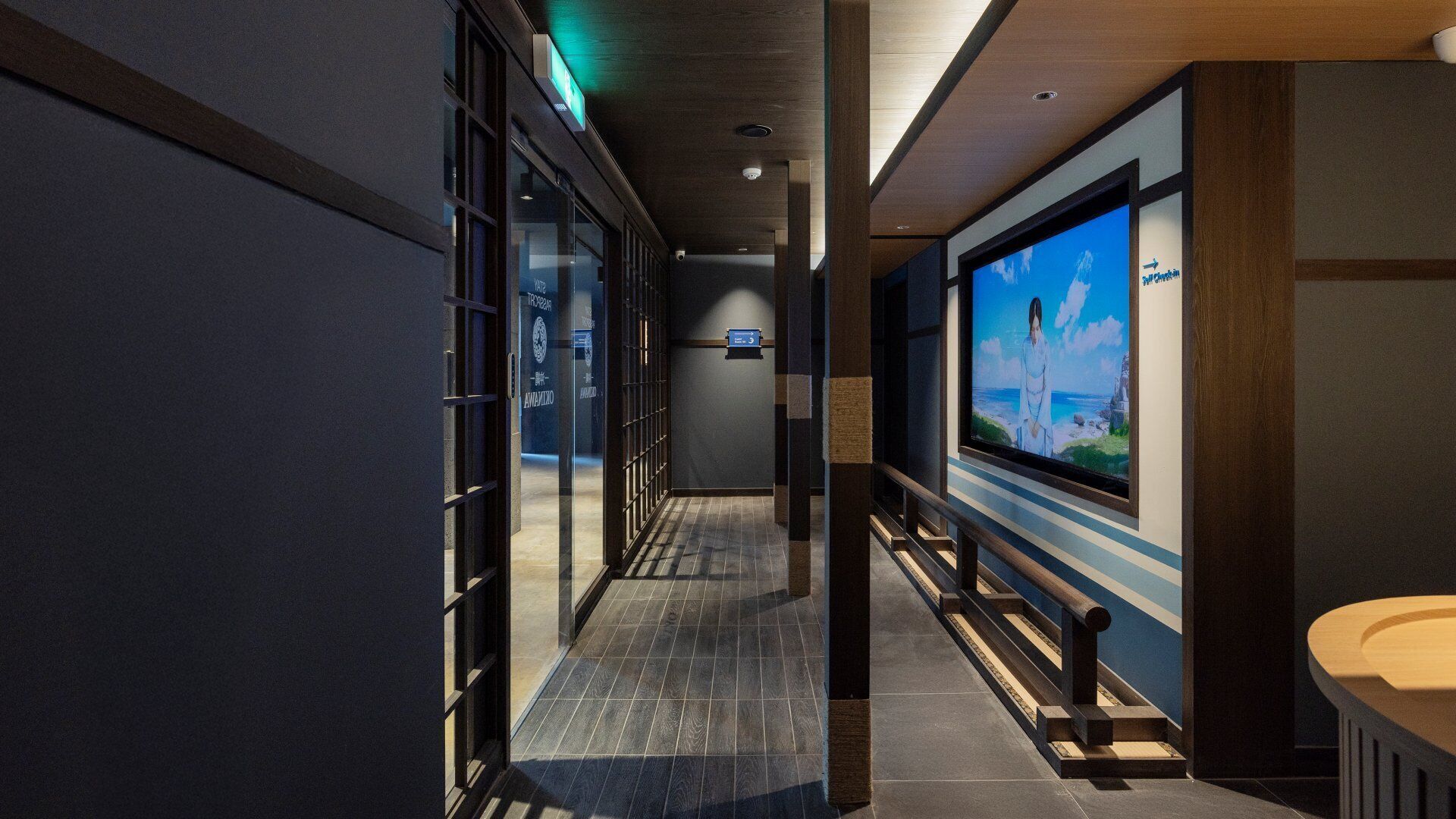 Un pasillo moderno de hotel con puertas de vidrio, un banco de madera y una gran pantalla de televisión que muestra una imagen panorámica.