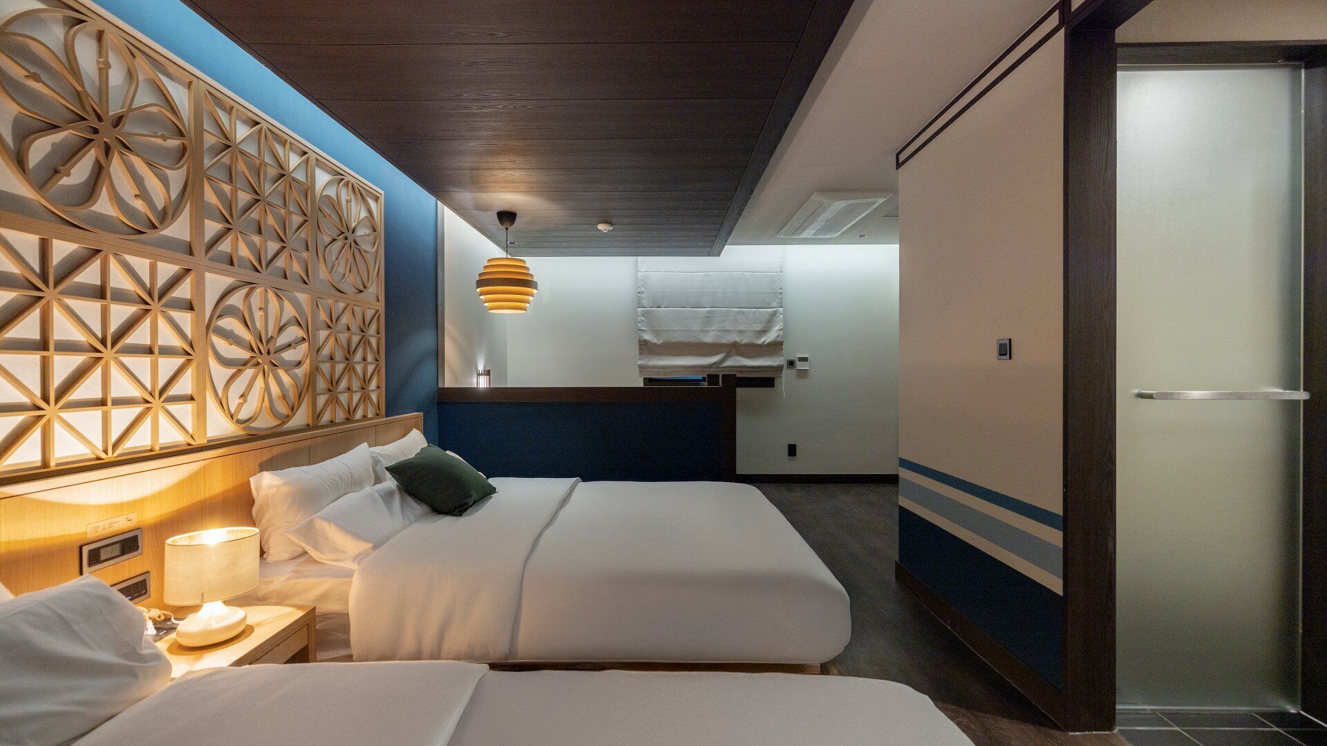Habitación de hotel con dos camas, un panel decorativo en la pared, una lámpara de noche y una lámpara colgante.