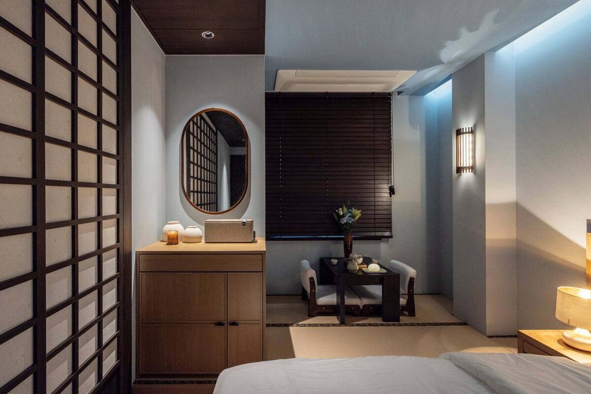 A modern hotel room with a bed, a wooden dresser, a desk with a chair, a mirror, and a window with blinds.