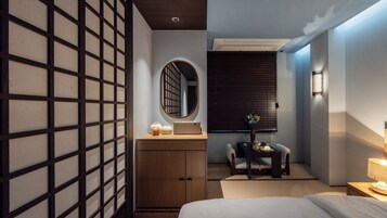 A modern hotel room with a bed, a wooden dresser, a desk with a chair, a mirror, and a window with blinds.
