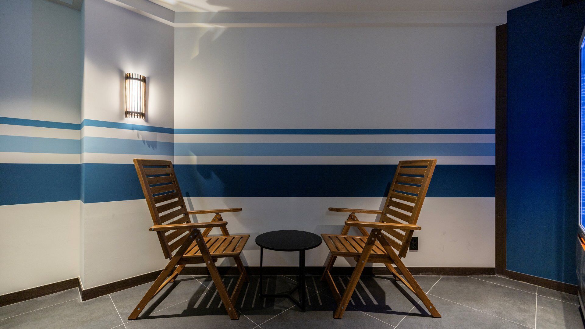 A corner room with two wooden chairs and a small round table, a wall with blue stripes, and a light fixture.