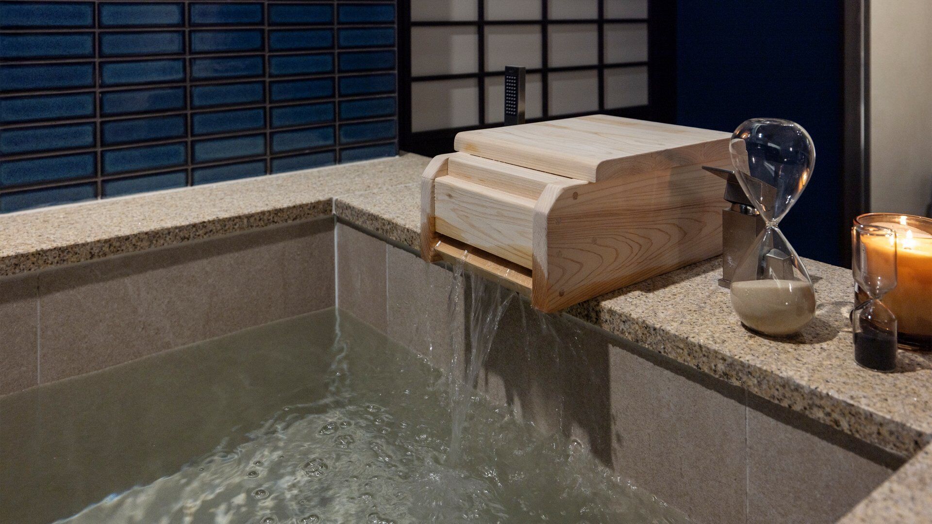 A bathtub with a wooden box and an hourglass on the edge, a candle, and a tiled wall in the background.