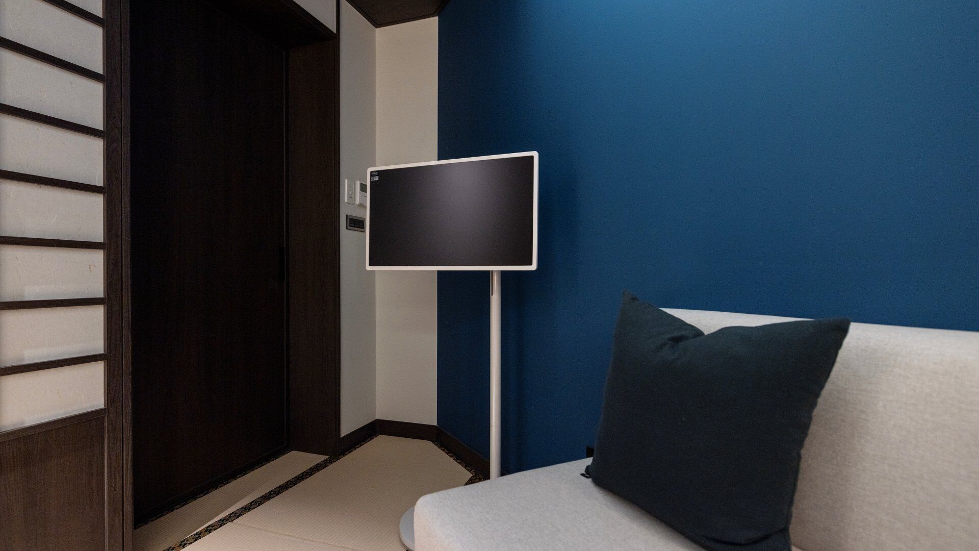 A hotel room with a dark wooden door, a blue accent wall, a white sofa with a dark pillow, and a flat-screen TV mounted on a stand.