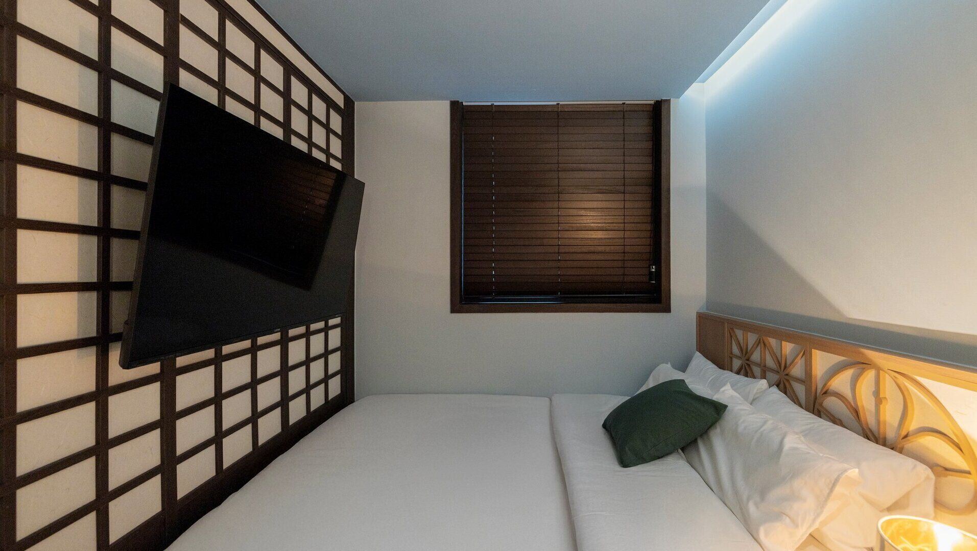 A bedroom with a bed, a TV mounted on a wall, a window with blinds, and a headboard with a decorative design.