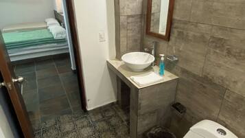 A bathroom with a white sink, mirror, and toilet. There is a small trash bin and a bottle of hand soap on the sink. The floor is tiled with a mosaic pattern.