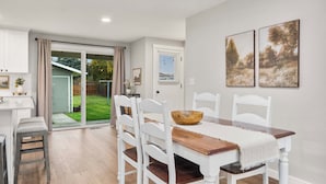 Un coin repas avec une table en bois, des chaises blanches et un saladier. Une porte-fenêtre coulissante donne accès à un jardin.