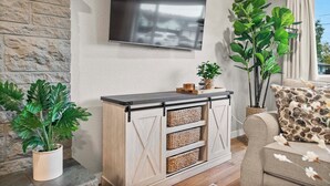 Un salon avec un mur en pierre, un téléviseur fixé au mur, une console en bois avec des paniers, un canapé et de grandes plantes vertes.
