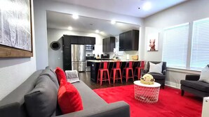 Una sala de estar moderna con un sofá gris, detalles rojos, un tapete rojo y una cocina con armarios oscuros y electrodomésticos de acero inoxidable.