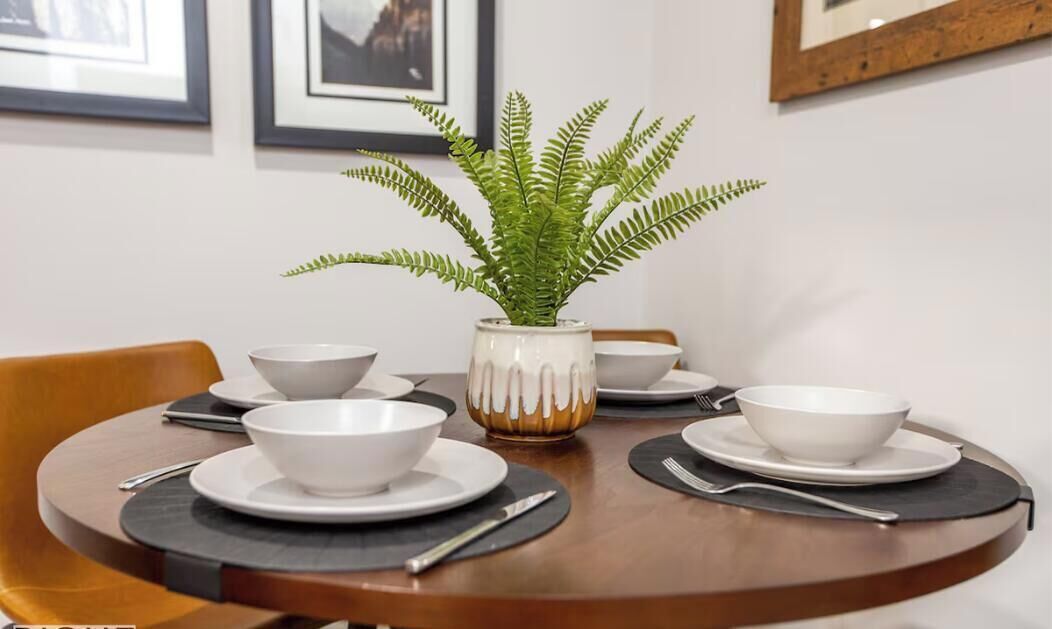 Une table à manger avec des bols, des assiettes et des couverts blancs, un vase contenant une plante verte et des tableaux encadrés accrochés au mur.