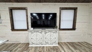 Una habitación con techo de madera, dos ventanas con persianas, un televisor de pantalla plana sobre un mueble blanco, y un suelo de madera con patrón de espiga.