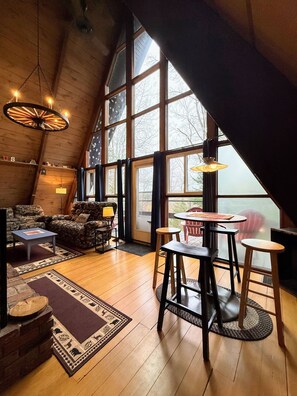 Une pièce confortable de style loft, avec un plafond haut, des poutres en bois, une grande fenêtre, une cheminée et un coin repas équipé de tabourets.